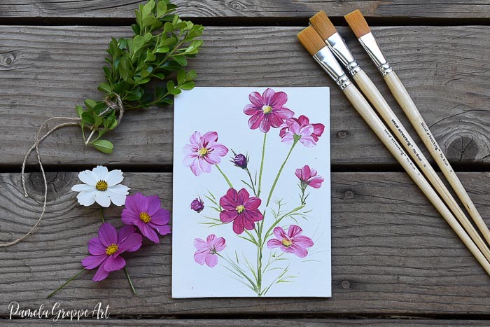 painting of cosmos flowers with brushes, boxwood cuttings and real cosmos flowers on wood surface