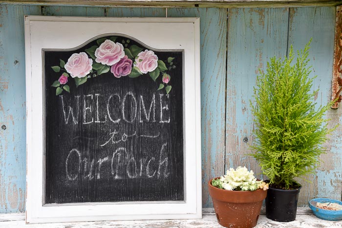 cabinet door made into a chalkboard sign. Outer frame is white with black chalkboard on the interior, hand painted roses arch over the top and chalk lettering reads, welcome to our porch.