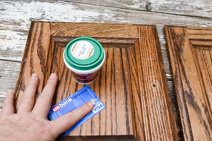 cabinet door laying on a table with a tub of wood filler and a credit card on top