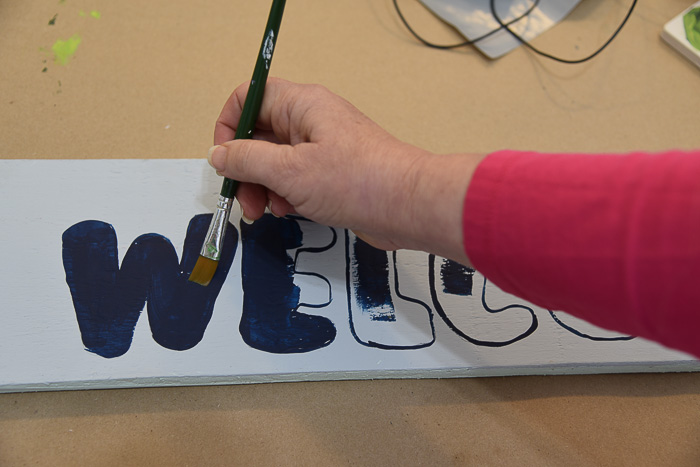woman painting in a w letter with dark blue paint on a gray painted board to make a welcome sign